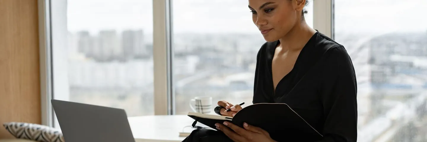 An actor sits at a desk at home, working on a laptop beside a notebook.